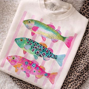 May include: Cream-colored t-shirt featuring three colorful fish designs. The fish have vibrant hues of pink, green, blue, and yellow, with decorative patterns. Pink vertical stripes are in the background. The shirt is folded on a patterned surface.