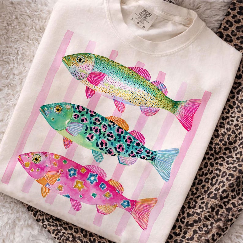 May include: Cream-colored t-shirt featuring three colorful fish designs. The fish have vibrant hues of pink, green, blue, and yellow, with decorative patterns. Pink vertical stripes are in the background. The shirt is folded on a patterned surface.