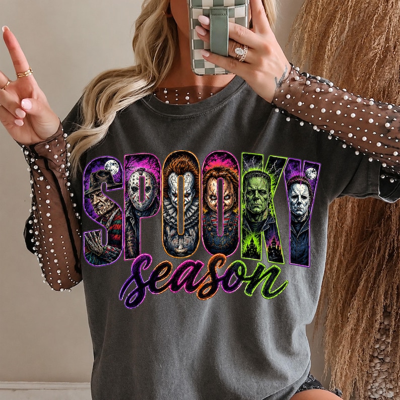 May include: A charcoal grey t-shirt featuring long sheer sleeves adorned with small white pearls. The front displays the text "SPOOKY season" in vibrant colours, with each letter containing a detailed illustration of a classic horror movie character.