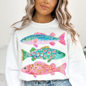 May include: White sweatshirt featuring three colorful fish illustrations. The fish have blue, green, and pink bodies with gold and pink accents. The sweatshirt is a casual, comfortable style.
