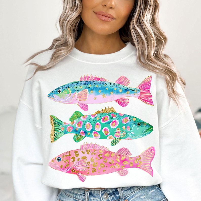 May include: White sweatshirt featuring three colorful fish illustrations. The fish have blue, green, and pink bodies with gold and pink accents. The sweatshirt is a casual, comfortable style.