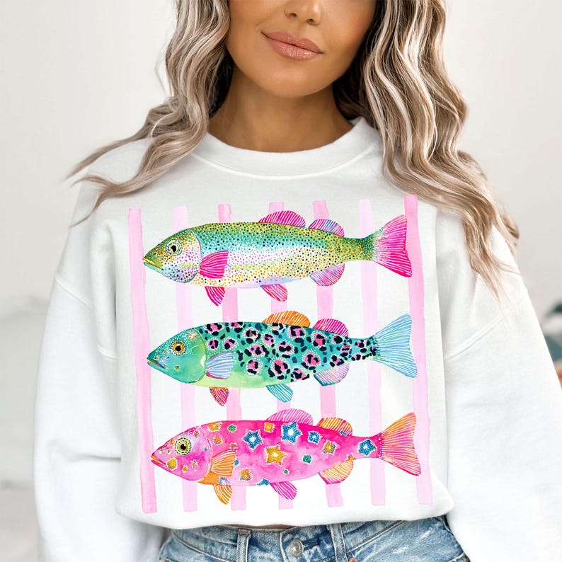 May include: White sweatshirt featuring three colorful fish designs. The fish are in shades of green, blue, and pink, with intricate patterns and pink fins. Vertical pink stripes are behind the fish.