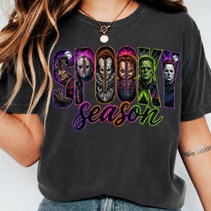 May include: A charcoal grey t-shirt featuring a graphic print with the text SPOOKY in large block letters, each containing a different horror movie character, and the word season in purple and green cursive script below the characters.