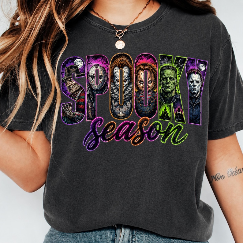 May include: A charcoal grey t-shirt featuring a graphic print with the text SPOOKY in large block letters, each containing a different horror movie character, and the word season in purple and green cursive script below the characters.