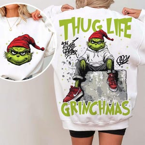 Puede incluir: Sudadera blanca con un gráfico del Grinch con un gorro de Papá Noel rojo. La parte posterior de la sudadera tiene un gráfico del Grinch con capucha, sentado en un bloque, con las palabras "THUG LIFE" y "GRINCHMAS" en verde.