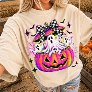 May include: A beige short-sleeve t-shirt featuring a graphic print of a vibrant pink and orange jack-o'-lantern filled with white ghosts. The pumpkin is topped with a large black and white checkered bow, surrounded by purple spiderwebs and bats.