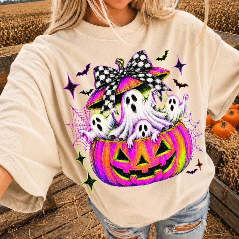 May include: A beige short-sleeve t-shirt featuring a graphic print of a vibrant pink and orange jack-o'-lantern filled with white ghosts. The pumpkin is topped with a large black and white checkered bow, surrounded by purple spiderwebs and bats.
