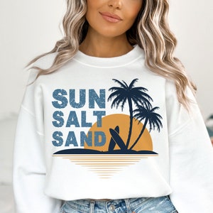 May include: White sweatshirt with a graphic design of palm trees, a sunset, and the text "SUN SALT SAND" in a distressed blue font.