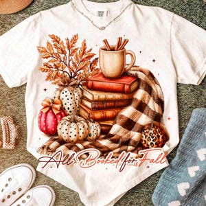 Peut inclure: Un t-shirt blanc &agrave; manches courtes avec une impression graphique repr&eacute;sentant une pile de trois livres, une tasse avec des b&acirc;tons de cannelle, des feuilles d'automne et trois citrouilles. Une couverture &agrave; carreaux marron et blanc est drap&eacute;e en dessous. Le texte indique All Booked for Fall.