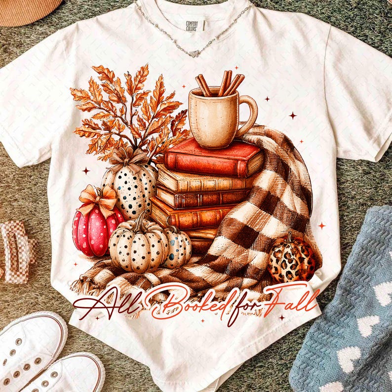 Peut inclure: Un t-shirt blanc &agrave; manches courtes avec une impression graphique repr&eacute;sentant une pile de trois livres, une tasse avec des b&acirc;tons de cannelle, des feuilles d'automne et trois citrouilles. Une couverture &agrave; carreaux marron et blanc est drap&eacute;e en dessous. Le texte indique All Booked for Fall.