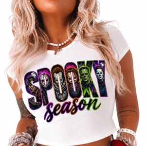 May include: A white short-sleeved t-shirt featuring a graphic print with the text "SPOOKY season" in bold, colourful lettering. The letters contain illustrations of various horror movie characters, including Freddy Krueger, Jason Voorhees, Pennywise, Chucky, Frankenstein's monster, and Michael Myers.