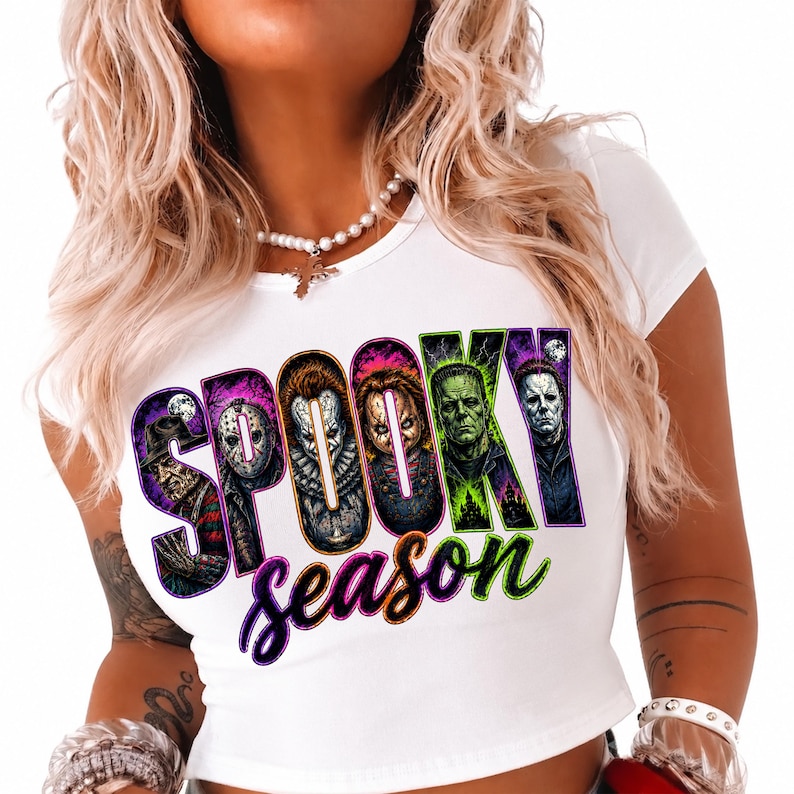 May include: A white short-sleeved t-shirt featuring a graphic print with the text "SPOOKY season" in bold, colourful lettering. The letters contain illustrations of various horror movie characters, including Freddy Krueger, Jason Voorhees, Pennywise, Chucky, Frankenstein's monster, and Michael Myers.