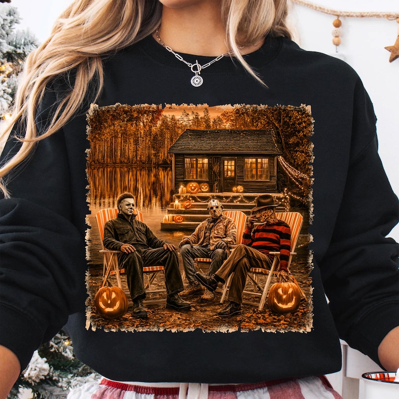 May include: A black sweatshirt featuring a graphic print of three horror movie characters sitting in lawn chairs in front of a cabin by a lake at night. The scene includes glowing jack-o'-lanterns, autumn trees, and warm cabin lighting.