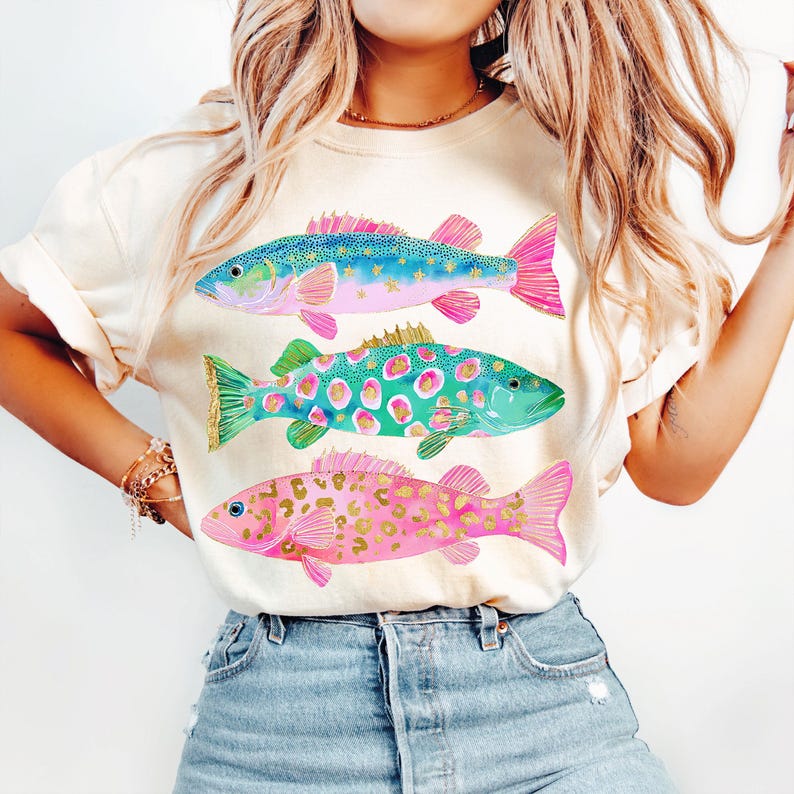May include: Cream-colored t-shirt featuring three colorful fish illustrations. The fish are in shades of blue, green, and pink, with gold and leopard print accents. The design is a whimsical and artistic representation of aquatic life.