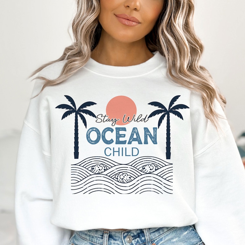 May include: White sweatshirt with a graphic design of palm trees, a sun, waves, and the text "Stay Wild Ocean Child".
