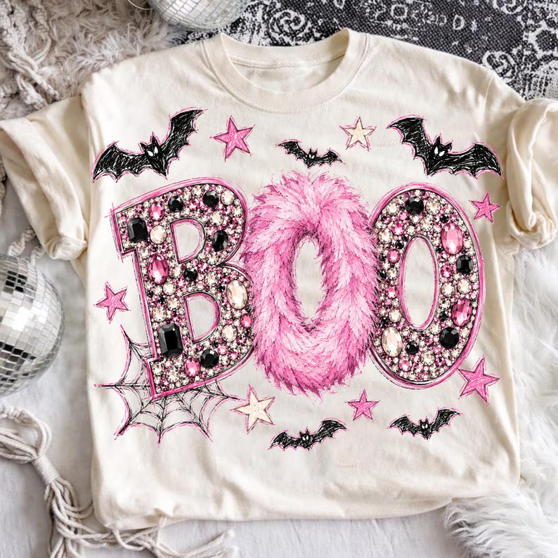 Op de afbeelding: Een cr&egrave;mekleurig t-shirt met het woord BOO in grote letters. De B en de O zijn gevuld met roze en zwarte strass-steentjes, terwijl de middelste O is gemaakt van roze imitatiebont. Het ontwerp bevat zwarte vleermuizen, roze sterren en een spinnenweb.