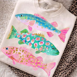 May include: Cream-colored sweatshirt featuring three colorful fish designs. The fish have blue, green, and pink scales with gold accents. The fish are arranged vertically on the shirt, with a leopard print pattern visible in the background.