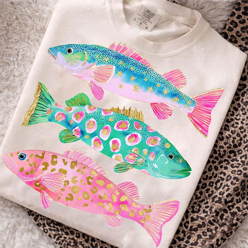 May include: Cream-colored sweatshirt featuring three colorful fish designs. The fish have blue, green, and pink scales with gold accents. The fish are arranged vertically on the shirt, with a leopard print pattern visible in the background.
