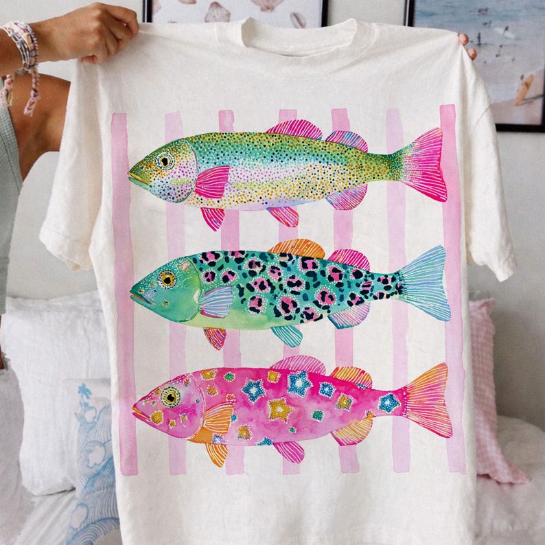 May include: White t-shirt featuring three colorful fish designs. The fish have vibrant patterns and colors, including pink, blue, green, and yellow. Vertical pink stripes are behind the fish. The shirt is held up by a person.