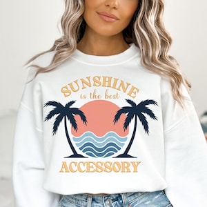 May include: White sweatshirt with a graphic design of a sunset with palm trees and the text "Sunshine is the best accessory".