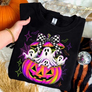May include: A black crewneck sweatshirt featuring a graphic of a vibrant orange and pink jack-o'-lantern with a black and white checkered bow on top. Three white ghosts emerge from the pumpkin, surrounded by purple spiderwebs, bats, and stars.
