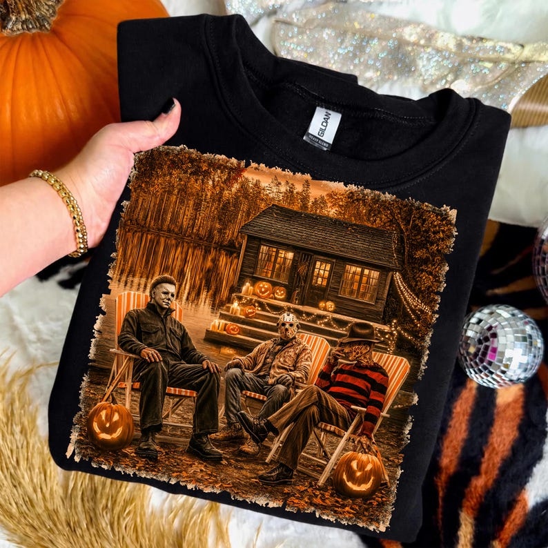 May include: A black crewneck sweatshirt featuring a graphic print of three horror movie characters sitting in lawn chairs in front of a cabin at night. The scene includes glowing jack-o'-lanterns, a dark forest, and a lake reflecting the cabin lights.