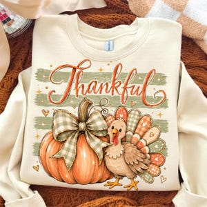 May include: A cream-colored crewneck sweatshirt featuring a graphic print of an orange pumpkin with a green and white gingham bow, a brown turkey with patterned tail feathers, and the word Thankful in orange cursive script with stars.