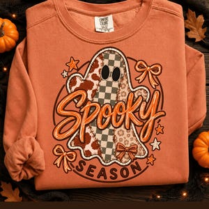 Peut inclure: Un sweat-shirt à col rond couleur terre cuite avec un motif central représentant un fantôme à carreaux et imprimé animal. Le fantôme est entouré d'étoiles et de nœuds, avec le texte orange "Spooky SEASON" en écriture cursive et en lettres capitales.