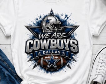 May include: A white short-sleeve t-shirt featuring a graphic print of a silver football helmet, a brown football, and a blue star. The text reads WE ARE COWBOYS DALLAS in bold silver and blue lettering. The design includes blue paint splatter accents.