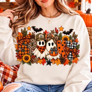 May include: A cream-coloured sweatshirt featuring a graphic print of two ghosts with the text SPOOKY. The design includes orange and black plaid patterns, sunflowers, autumn leaves, bats, stars, a pumpkin, and a camouflage cap with the word BOO.