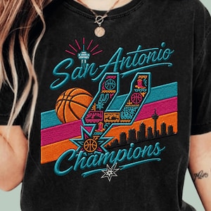 May include: A black t-shirt featuring a graphic design with the text San Antonio Champions. The design includes a basketball, a city skyline silhouette, and a large stylized letter U filled with colorful icons like boots and basketballs.