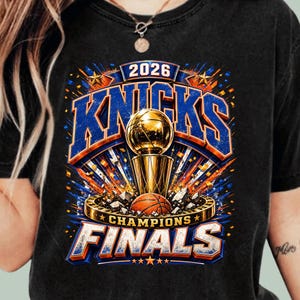 May include: A black t-shirt featuring a graphic print with the text 2026 KNICKS CHAMPIONS FINALS. The design includes a gold basketball trophy, a basketball, blue and orange accents, and star motifs, creating a vibrant sports-themed illustration.