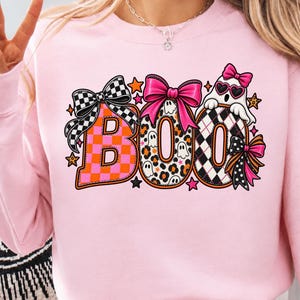 May include: A light pink sweatshirt featuring a graphic print of the word BOO in large letters. The B is orange and pink checkered with a black and white checkered bow. The O is leopard print with a pink bow. The final O is black and white plaid.