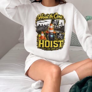 Op de afbeelding: Witte sweatshirt met een geel en zwart grafisch ontwerp met de tekst "Hoist the Cone", verkeerskegels, een lasmasker en een doodshoofd. Het ontwerp bevat de tekst "EST. 2025".