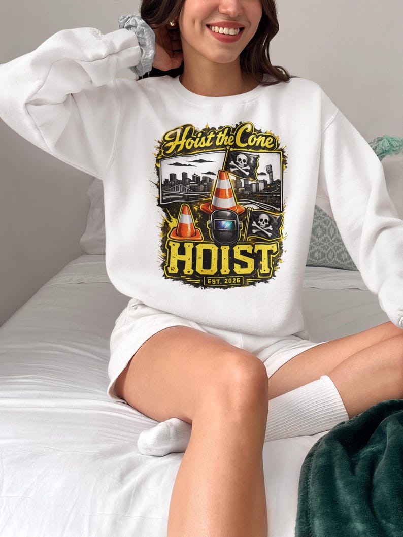 Op de afbeelding: Witte sweatshirt met een geel en zwart grafisch ontwerp met de tekst "Hoist the Cone", verkeerskegels, een lasmasker en een doodshoofd. Het ontwerp bevat de tekst "EST. 2025".