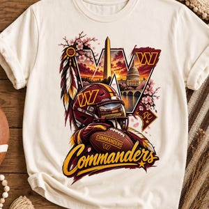 May include: A cream-coloured short-sleeve t-shirt featuring a graphic of a Washington Commanders football helmet, a football, the Washington Monument, the U.S. Capitol, cherry blossoms, and feathers. The text reads Commanders, W, and DC.