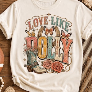 Könnte beinhalten: Ein cremefarbenes T-Shirt mit einem Grafikdruck und dem Text LOVE LIKE DOLLY. Das Design zeigt einen türkisfarbenen Cowboystiefel gefüllt mit Blumen, zwei rosa Rosen, drei Schmetterlinge und ein Hufeisen, alles in einer farbenfrohen Gestaltung.