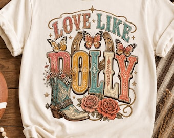 Può includere: Una t-shirt color crema con una stampa grafica che riporta il testo LOVE LIKE DOLLY. Il design include uno stivale da cowboy turchese riempito di fiori, due rose rosa, tre farfalle e un ferro di cavallo, il tutto in una tavolozza colorata.