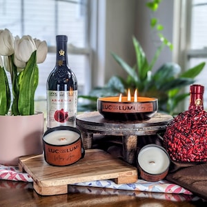 May include: A still life arrangement featuring candles, wine, and decorative elements. A large black candle with three wicks is lit, alongside smaller candles and a bottle of wine. White tulips in a pink pot and a decorative red bottle are also present.