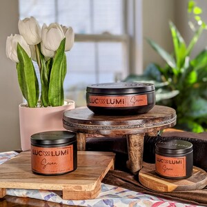 May include: Black candle tins with the text "Luc Lumi" and "Sevan" on brown labels. The candles are displayed on wooden risers and a wooden tray. White tulips in a pink pot are in the background.