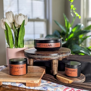 May include: Several black candles with brown labels and the text "LUC LUMI" and "Apricot" are displayed on wooden stands. A bouquet of white tulips in a pink pot and a green plant are in the background. The candles are of varying sizes.