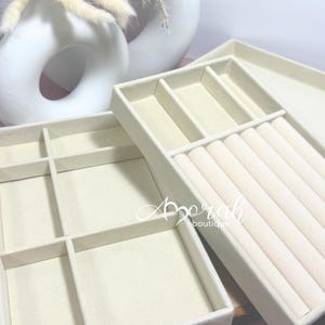 May include: Beige jewelry organizer trays with various compartments for rings, necklaces, and other accessories. The trays are made of a soft, felt-like material and are designed to protect jewelry. The trays are stacked on top of each other.