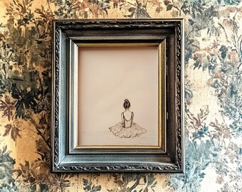 May include: A framed black and white drawing of a ballerina in a tutu, sitting with her back to the viewer. The drawing is on a light beige background and is framed in a dark gray frame with a decorative border.