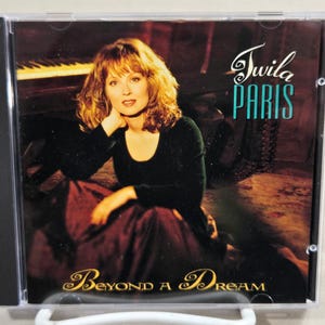 May include: A compact disc case with a photo of Twila Paris. The case is black with a clear plastic front. The photo shows Twila Paris sitting on a piano bench, wearing a black shirt and a burgundy skirt. The text on the case reads "Twila Paris" and "Beyond a Dream".