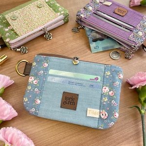 Op de afbeelding: Verschillende stoffen portemonnees in diverse kleuren en bloempatronen worden getoond. De portemonnees hebben ritsen, kaartsleuven en decoratieve bedels. Eén portemonnee is lichtblauw met een bloemenontwerp en een bruin leren label met "torie & ren".