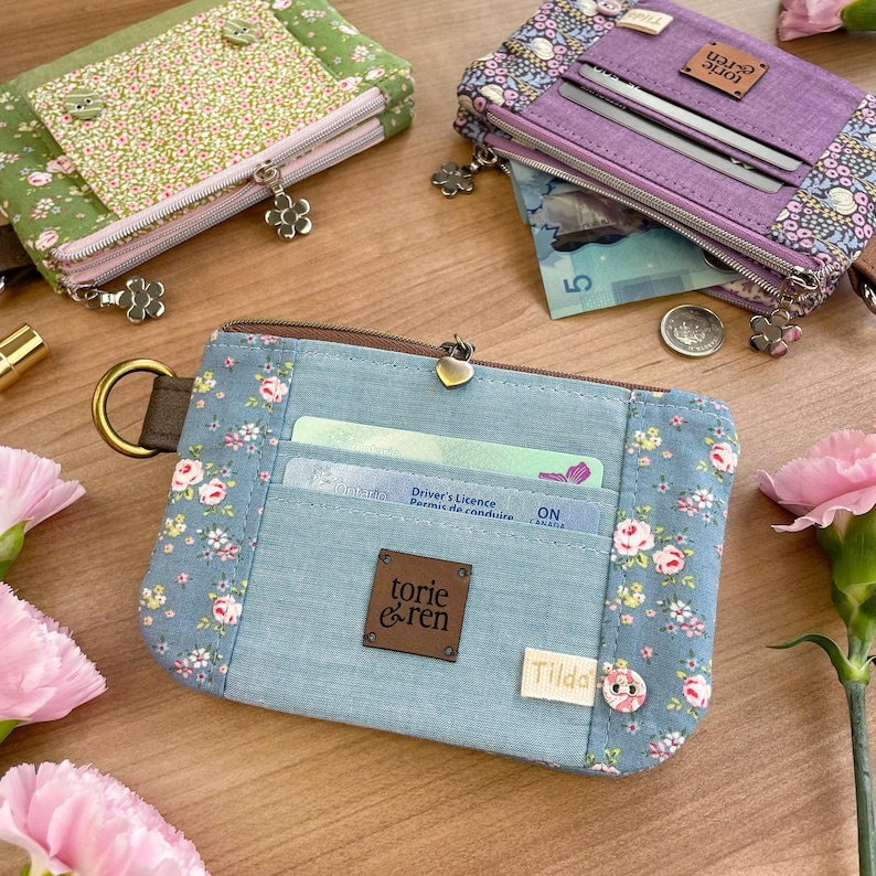 May include: Several fabric wallets in various colors and floral patterns are displayed. The wallets have zippers, card slots, and decorative charms. One wallet is light blue with a floral design and a brown leather tag that says "torie & ren".