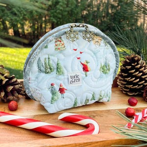 Op de afbeelding: Een blauwe, gewatteerde toilettas met rits en een winterse print. De tas heeft een sneeuwvlok-bedel en de tekst "torie & ren".