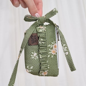 May include: A small olive green zippered pouch featuring a white polka dot pattern and a floral print with light pink flowers. The pouch has a ruffled fabric trim, a tied bow at the top, and a brown rectangular tag with the text torie & ren.