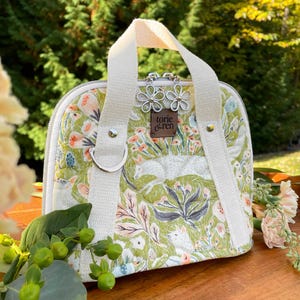 May include: A small, quilted handbag with a floral and fox pattern in green, white, and peach. It has a white handle and straps, silver flower charms, and a small leather tag that reads "torie & ren".