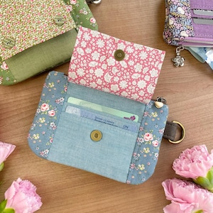 May include: Floral print wallets in shades of pink, blue, and green. The open wallet reveals a driver's license and credit cards. The wallets have a snap closure and a metal ring for attaching keys. The overall theme is a floral pattern.
